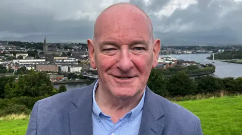 Mark Durkan, pictured from the chest up, wearing a blue shirt and grey suit jacket. He is bald. 