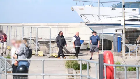 National Crime Agency Raymond Nuttall, Colin Benson and Craig Nuttal walk through the marina car park in Watchet on a sunny day.