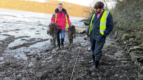 A person with a red top holding a buoy and seaweed and another person with a hi vis and blue overalls on a muddy foreshore