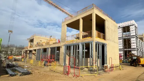 A building site showing large constructions which are just concrete and metal shells. There are safety barriers around the constructions site and the arm of a yellow crane can be seen overhead.