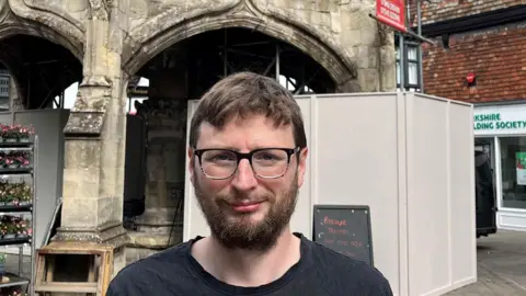Sam Charleston standing in front of the Poultry Cross monument. He is wearing a dark T-shirt and has brown hair and glasses. Hoarding has been placed around the stone monument.