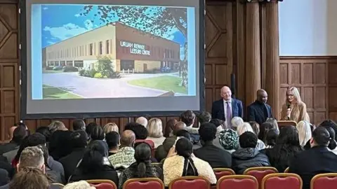 Sheffield City Council The ceremony in Sheffield Town Hall, where an audience is sat in front of a projector screen, showing a mock-up of the new leisure centre with Uriah Rennie's name on it.