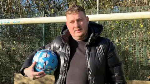 Charlie Barker is standing in front of a football goal. He is wearing a black t-shirt, black puffer jacket and is holding a blue football under one arm.