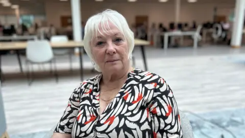 Kate Bradbrook/ BBC Woman in a white and black dress sitting in an office