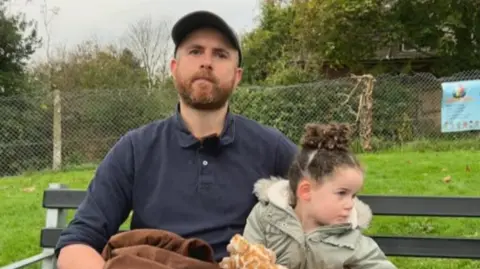 Martin Reid has a ginger beard, a navy, long-sleeve polo and a light green hat. He is sitting down on a bench and looking at the camera next to his daughter, who is wearing a grey coat and has her curly hair tied up.