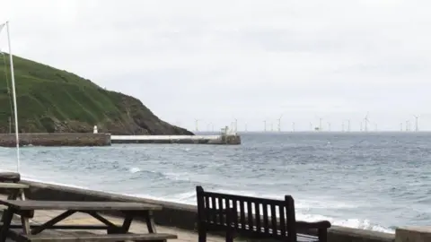 Orsted Laxey beach with the tide in from the promenade, with a wooden bench to the right. There is a green headland to the left above a stone pier. On the horizon are dozens of wind farm turbines. It is a grey day.