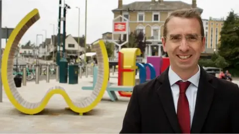 Peter Taylor Peter smiles at the camera while standing outside Watford Station and in front of play equipment. he wears glasses and a suit.