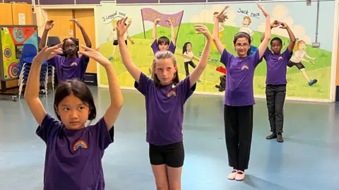 BBC Five pupils from Stanton Bridge Primary School in a ballet pose, in their sports hall. They're wearing purple tshirts. 
