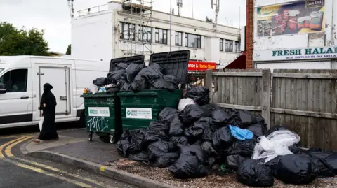 PA Media A woman dressed in a black face covering walks past over flowing green bins with black back piled up high.