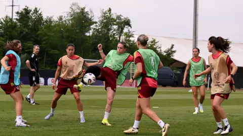 Six members of the Wales womens football team in the middle of training. The are on a football pitch with a referee in the back. Some are wearing green vests, blue and orange vests. 