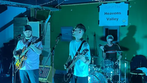 Heavens Valley Community Benefit Society A band, consisting of two men on guitars and a third on drums sat behind them, performing in a green-lit spot