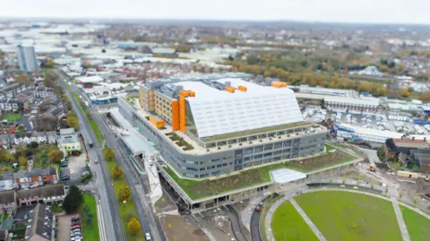 Sandwell and West Birmingham Hospitals NHS Trust A aerial view of a new super hospital which has a white sloped roof and orange frame, with roads and a large grass area surrounding it