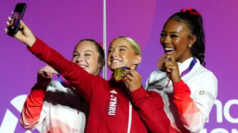Image of three teenagers wearing tracksuits and holding up medals as they take a selfie standing on a podium.