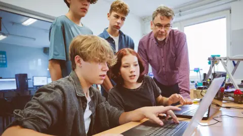 Two students seated at a desk, looking at a laptop with two others standing behind them as a teacher looks on, supervising