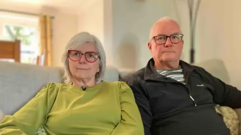 A middle aged man and woman sit next to eachother on the sofa. She is wearing a green top and red glasses, and he is wearing a dark fleece and glasses.