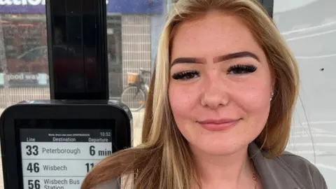 Laurisa Hallam who has long fair hair and is wearing a grey jacket. She is standing in a bus shelter and to her right is a black metal box which says the 33 to Peterborough is due to depart in six minutes. 