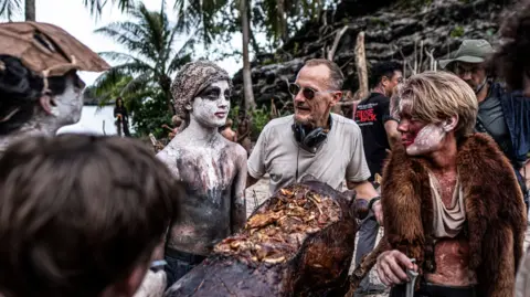 BBC/Eleven/J Redza Marc Munden, wearing sunglasses with headphones around his neck, speaks to two of the actors, who are in costume.