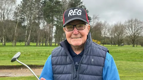 A man with black rimmed glasses is pictured smiling whilst holding a golf putter with a black and red cap featuring his name, Reg, in white capital letters. A golf course is in the background. He's also sporting a navy gilet over a light blue jumper and navy blue polo shirt. 