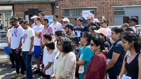 A crowd of people, some wearing sportswear, standing and looking solemn. There's a brick building behind them and it's a sunny day.