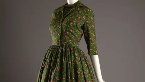 The Harris The torso and hips of a cream mannequin against a dark grey background. It is wearing a dark olive green dress with small red roses. The dress is cinched at the waist with a full skirt, has elbow-length sleeves and a v-neck with an insert in the same fabric.