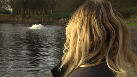 A woman who is a victim of crime who the BBC has agreed not to name, is looking away from the camera towards a water feature in a lake to avoid showing her face. She has blonde hair.