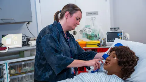 A woman in a dark blue shirt holds a medical device to the ear of a manikin lying in a hospital bed. They are surrounded by medical equipment