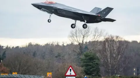 Getty Images Dark gray US fighter jet with US Air Force livery. It is coming in to land at an airbase. In the background trees can be seen. There is a traffic sign below it.