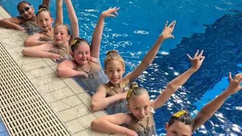 supplied Young girls perched on the side of a swimming pool, in the water, with arms raised.