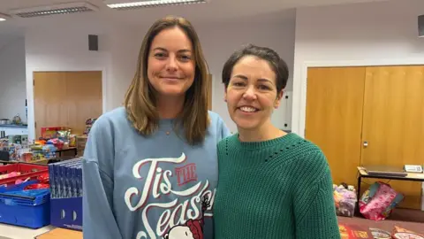 Two women stood side by side, looking into the camera and smiling. Laura, on the left, is wearing a blue jumper with 'Tis The Season' written on it in white letters. Penny, on the right, is wearing a dark green knitted jumper. They're pictured indoors, stood in front of rows of tables stacked high with crates full of food, toys, and toiletries.