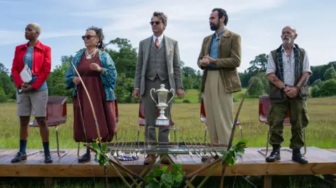 BBC Studios Five adults, two women and three men stand on a platform in a field in front of a makeshift table which on top sits a silver cup trophy in the middle, Either side of the trophy sit arrows for bows at either end of the table. The sky is bright with little cloud and the field is green with trees in the far background. The adults are looking to the left at something out of frame.
