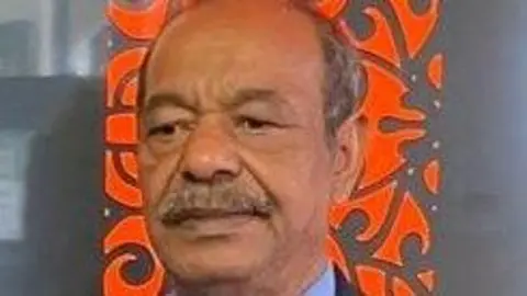 Abul Kalam has a moustache and is wearing a blue shirt and dark coloured suit jacket. He is standing in front of an orange patterned backdrop.