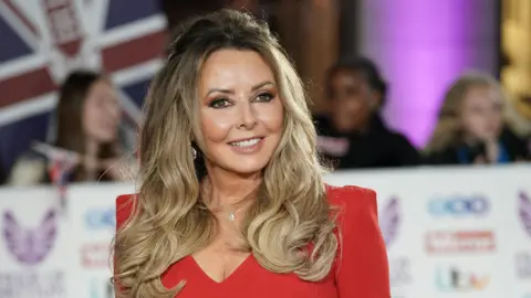 PA Media Carol Vorderman pictured at an event wearing a red v neck dress with shoulder pads. She has long blonde hair and is smiling. 