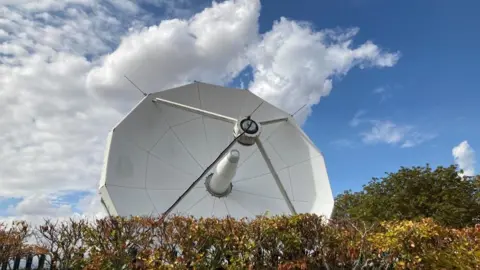 A large white satellite is pointing up to blue skies. 