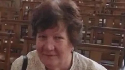 Family handout An image of Julie Bunkhall, smiling at the camera. She has short dark hair and is in a room with wooden seats behind her. She has on a light top with a strap on one shoulder. The image is slightly blurred.