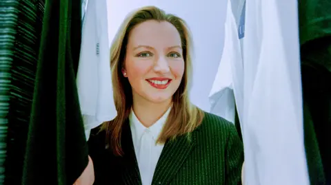 Wattie Chung Michelle Mone in a green pin-striped business jacket and white shirt in front of a rack of white shirts. She is smiling broadly and has long blonde hair.