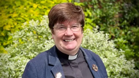 A mid-shot photo of Moira Astin. She has short brown hair, wears glasses and is smiling.