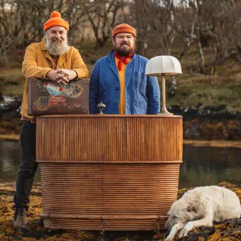 Shelley Richmond/Hello Halo/BBC Scotland Ro and Banjo stand behind what appears to be a reception desk - in bamboo rods in a rounded shape with a lamp on top and a reception bell. Both are wearing orange woolly hats and Granpa the dog is lying on the ground beside the desk.