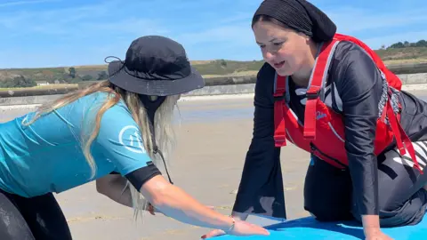 A Healing Waves volunteer is in a blue rash vest and black sun hat demonstrates a surfing technique on a blue surfboard laid on wet sand, while Maha is wearing a red life jacket and dark watersports clothing kneels on the board listening during a beach lesson. A clear blue sky and coastal dunes are visible in the background.