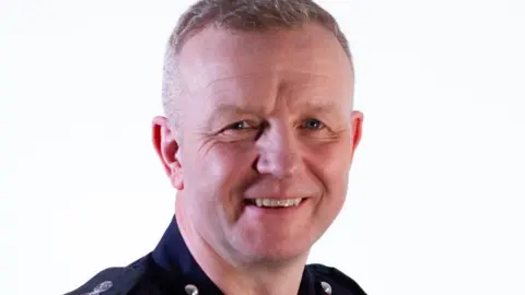 HIWFRS Neil Odin is wearing a blue uniform in front of a white background. He is smiling and has short, white hair 