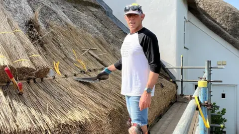 James Marshall James Marshall wearing a white T-shirt, blue jean shorts and a hat. He is stood by a thatched roof. 