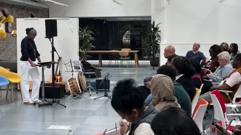 BSWN A black woman giving a talk in front of an audience in a large room. Musical instruments and microphones can be seen behind her.
