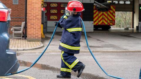 A child carrying a fire hose, dressed up as a firefighter