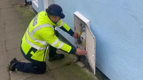 Wales & West Utilities The picture shows a person wearing a high-visibility jacket marked with the word “GAS” kneeling on a pavement next to a light blue building. The individual is working on an outdoor gas meter box, which has its door open to reveal the meter and connected pipework inside.