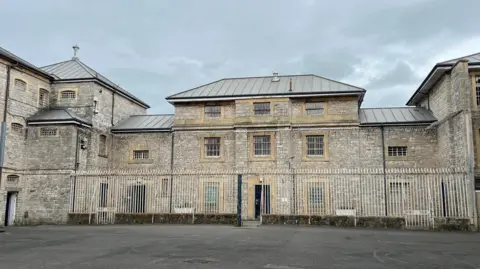 BBC A large grey stone prison building with white gates sits under a gloomy sky.