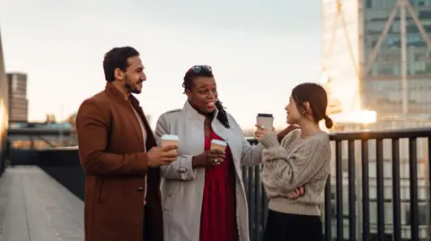 Getty Images A man and two women are standing outside in what looks to be London, it is at a time when the sun is going down - they sare standing close together and having a conversation. They appear to be business people on a break - they are all drinking coffee and holding cups 