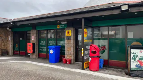 A picture of a garage. It is a brick and glass building. There is bins outside of it. The building is green and red in colour.
