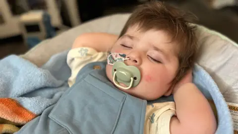Jack is asleep with his hands behind his head. He has brown hair, is in a blue baby grow and has a dummy in is mouth and a feeding tube going up his nose.