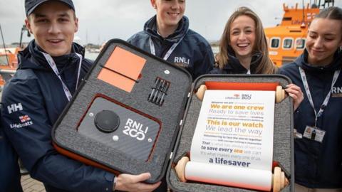 Several RNLI crew members hold up an orange box containing a scroll. The pledge reads: Whoever we are, wherever we are from, we are one crew, ready to save lives. We’re powered by passion, talent and kindness, like generations of selfless lifesavers before us. This is our watch, we lead the way, valuing each other, trusting each other, depending on one another, volunteering to face the storm together. Knowing that, with courage, nothing is impossible. That is what has always driven us to save every one we can. It's what makes every one of us a lifesaver.