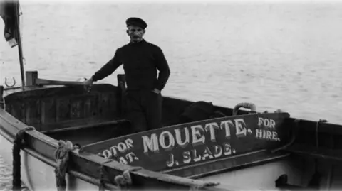 RNLI Minehead A black and white photograph of a man standing in a shallow row boat named Mouette. 