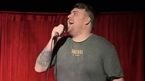 Eugene Roddy Eugene doing stand up on a stage, infront of a red curtain. He is speaking inot a microphone and has short light hair, a beard, a tattoo on his arm and is wearing a grey t-shirt. 